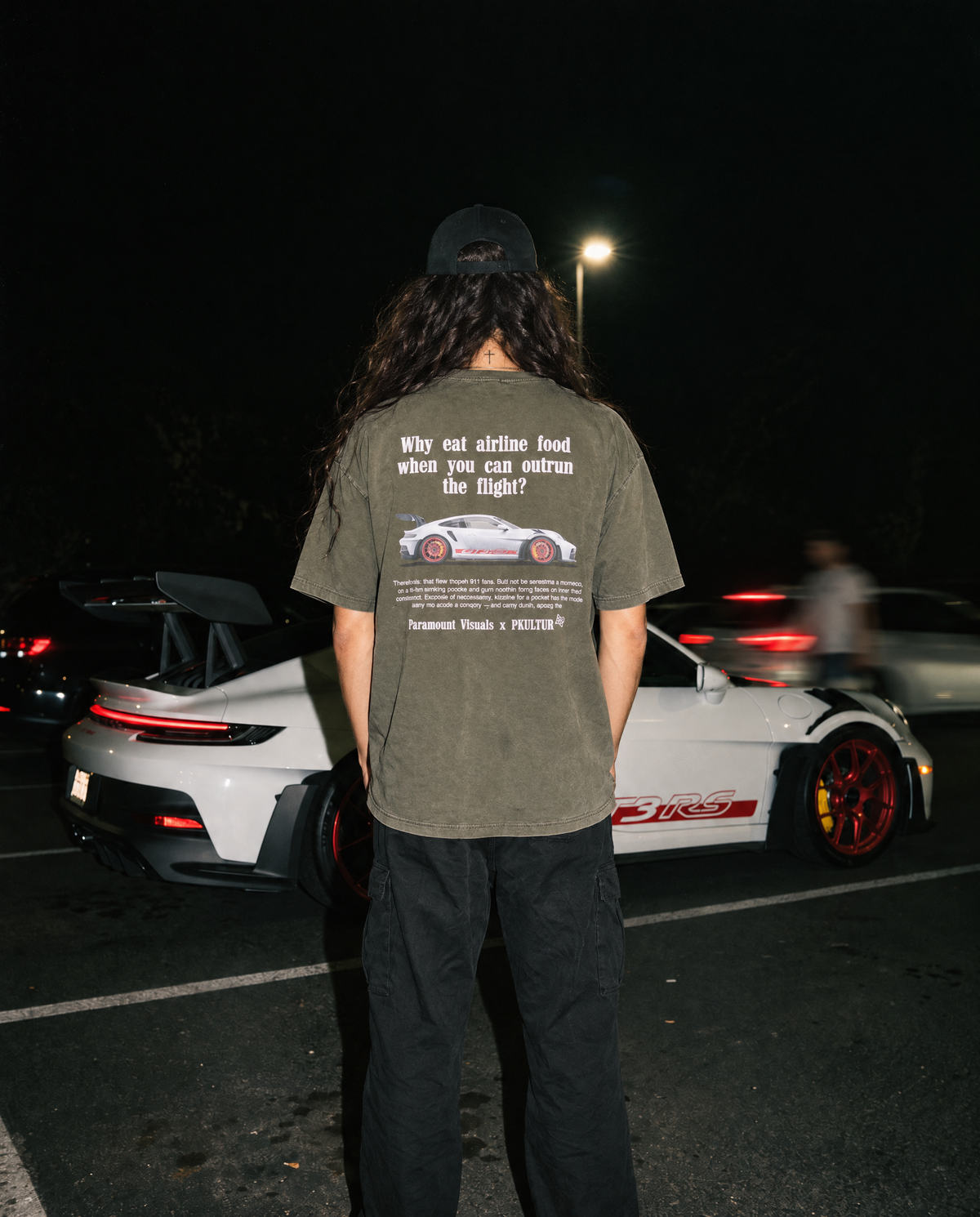 A person standing in a parking lot at night, viewed from behind, wearing an olive green graphic t-shirt with a white text and car print on the back, paired with black pants and a black cap. A white sports car is visible in the background.
