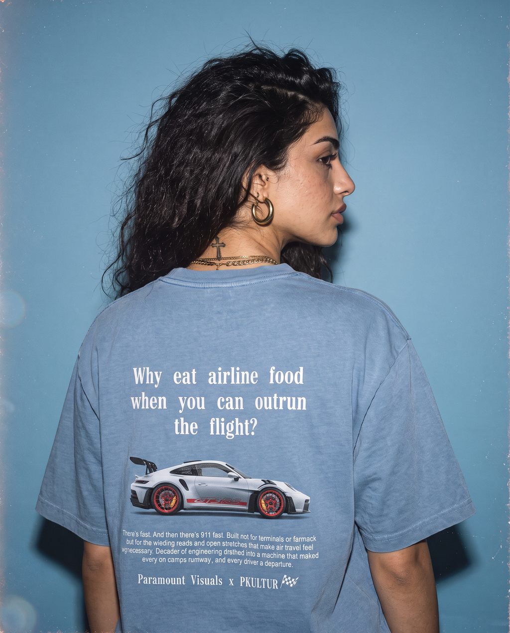 A woman shown from behind wearing a faded light blue graphic T-shirt against a blue background. The shirt features white text reading 'Why eat airline food when you can outrun the flight?' above a white sports car graphic and smaller text at the bottom.
